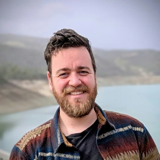 Markos smiling outdoors in a patterned jacket, with a hazy reservoir and hills behind him.